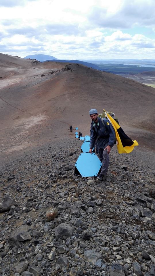 Figure carrying blue geometric form across volcanic ground, Námafjall