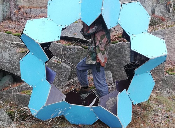 Figure inside a ring of light blue hexagonal panels among granite boulders, Borgåsen